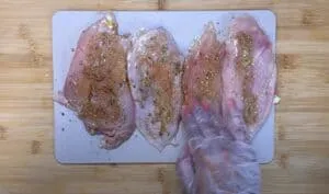 Raw chicken breasts seasoned with spices on a cutting board, with a gloved hand applying seasoning for cooking.